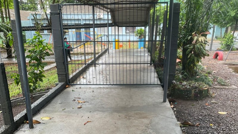 Em ambiente aberto, é mostrado o pátio da escola. Em primeiro plano, aparecem as grades do portão, de cor cinza escuro. Ao fundo, estão as paredes externas da escola, de cor azul piscina. Nos lados direito e esquerdo da foto aparecem plantas.