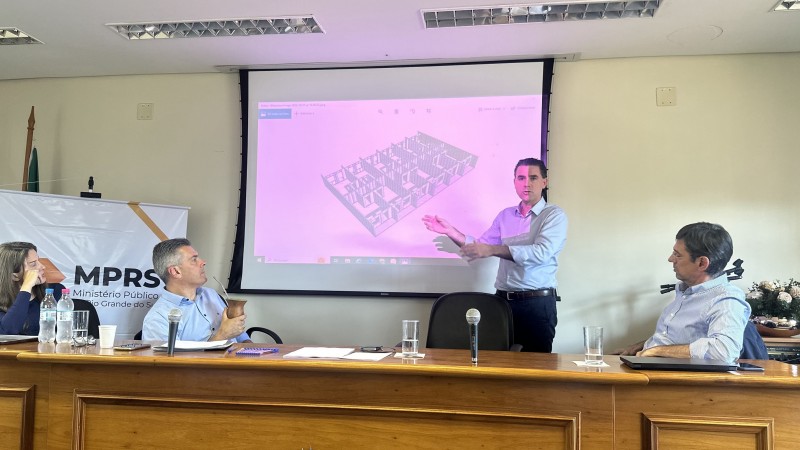 Num auditório, Peruchin está pé, apresentando um slide para a plateia, que não aparece na foto. À sua frente, há uma mesa junto à qual estão sentados dois homens e uma mulher, que fazem parte da reunião e acompanham a fala.