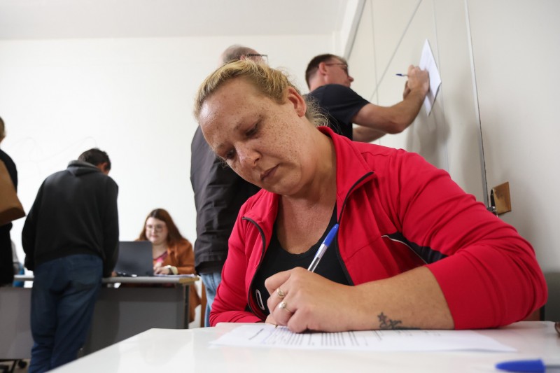 Foto em ambiente fechado. Uma mulher está sentada, debruçada sobre uma mesa, lendo e preenchendo a mão o recibo de doação e declaração de compromisso. Ao fundo, há outras pessoas desfocadas, sendo atendidas e assinando o documento.