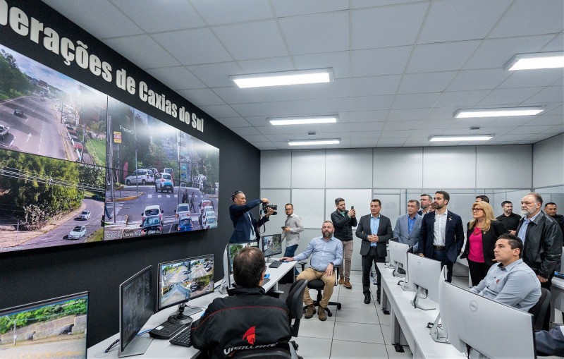 Eduardo Leite visita a sala do Centro de Monitoramento de Caxias do Sul. Ele está ao fundo e a direita acompanhado de diversa pessoas. À esquerda da imagem há uma grade tela onde se vê movimentação de carros em ruas diferentes. Há ainda duas grande bancadas com computadores e três homens a sentados diante de três desses computadores.