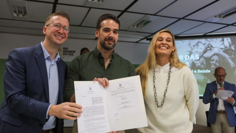 Foto posada em ambiente fechado. Fantinel, Leite e Helena Casanovas estão de pé e sorriem para a câmera. Os dois primeiros exibem o documento assinado.