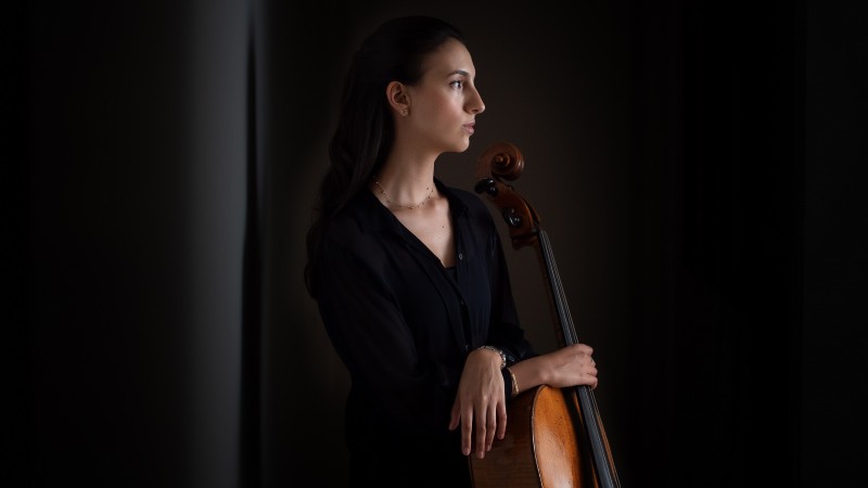 Foto de perfil da violoncelista Marina Martins. O olhar da artista está direcionado para a direita da imagem e ela segura seu violoncelo. A artista veste uma camisa preta e o fundo da imagem também é escuro, contrastando com a pele branca e com a cor marrom clara do instrumento musical. 