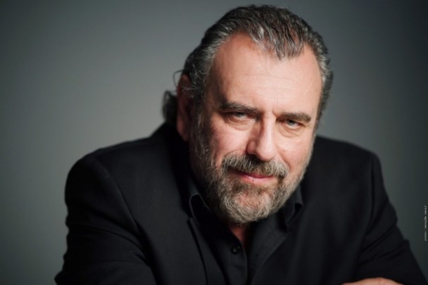 Retrato frontal de Luis Gorelik, maestro argentino, que encara a câmera. Homem de meia idade, com barba e cabelos grisalhos. O terno preto e o fundo escuro da fotografia contrastam com sua pele e olhos claros.