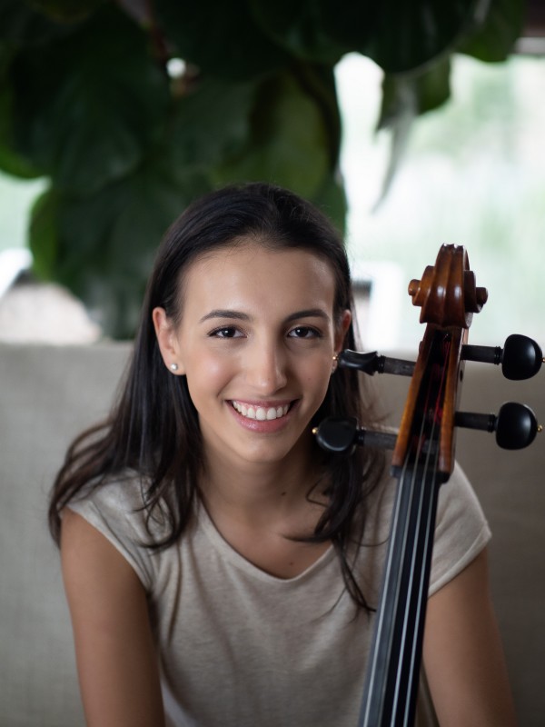 Retrato frontal de Marina Martins. A jovem sorri e olha para a câmera, segurando um violoncelo. Ao fundo, desfocadas, estão algumas folhas, formando um fundo verde e branco para o retrato.