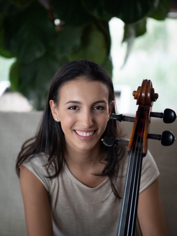 Retrato frontal de Marina Martins. A jovem sorri e olha para a câmera, segurando um violoncelo. Ao fundo, desfocadas, estão algumas folhas, formando um fundo verde e branco para o retrato. 