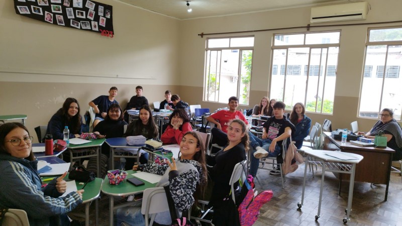Imagem de diversos alunos em suas carteiras em uma sala de aula. Mais à direita está a professora, também sentada à sua mesa.