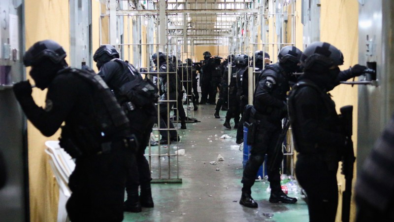 Imagem de um corredor de penitenciária com policiais armados diante das portas das celas fazendo a verificação no interior delas. 