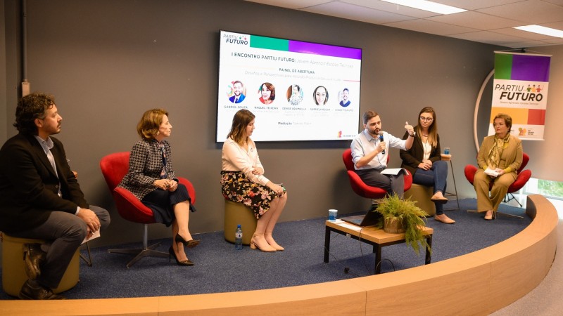 Em um pequeno tablado a seis pessoas sentadas em poltronas. Atrás delas há uma ela que exibe as fotos dos participantes do painel. Na quarta posição da esquerda para direita está o vice-governador Gabriel Souza com o Microfone nas mãos e gesticulando enquanto fala para a plateia.