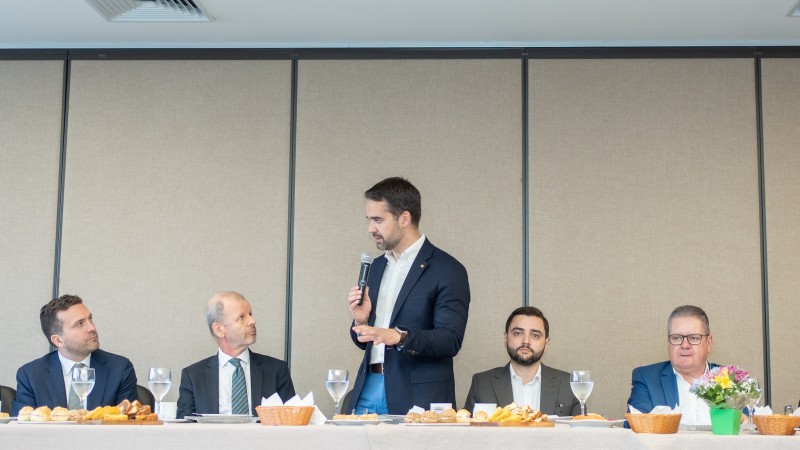 Foto frontal de uma mesa de café da manhã junto à qual estão Johannes Zutt, Eduardo Leite, Gabriel Souza e Ranolfo Vieira Junior. Leite, ao centro, está de pé, falando ao microfone e gesticulando com uma das mãos. Ele está levemente de lado