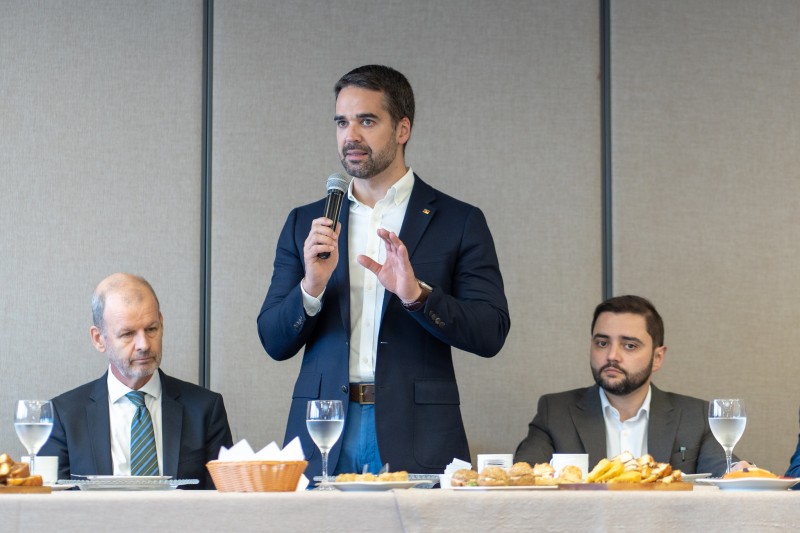 Foto frontal de Leite de pé, falando ao microfone na sala de reuniões. Ele está junto a uma mesa de café da manhã. A seu lado direito está Zutt; à esquerda do governador, está Gabriel Souza. Ambos estão sentados.