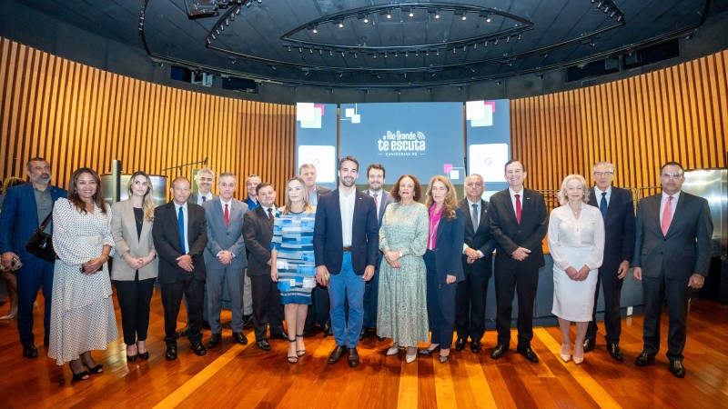 Foto posada, frontal, de corpo inteiro dos participantes do evento. No palco de piso amadeirado, Leite est&aacute; de p&eacute; junto a secret&aacute;rios e outras autoridades. Ele est&aacute; no centro da foto e sorri. Ao fundo, h&aacute; um tel&atilde;o com a logo do Rio Grande te escuta, contra um fundo azul. As paredes do fundo s&atilde;o de cor marrom e apresentam relevos.