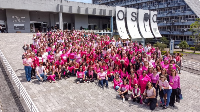 Foto posada em plano aberto de quase 300 pessoas, em ambiente aberto, vestidas em diferentes tons de rosa. Elas estão na rampa de cor cinza, em frente à Sedac e do letreiro da Ospa, tudo em fachada cinza. Há pessoas em pé, sentadas ou agachadas. A maior parte olha para a câmera e sorri