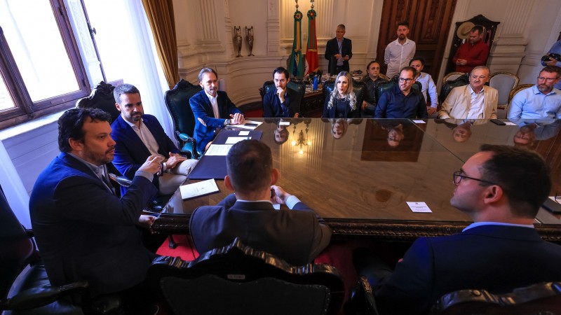 Foto geral de uma sala de reunião no Palácio Piratini. Os participantes estão sentados junto a uma grande mesa retangular de madeira. A imagem enquadra apenas uma parte dela. À esquerda, Leite, Lemos e Costella aparecem sentados. Ao redor da mesa, os prefeitos também se encontram sentados.