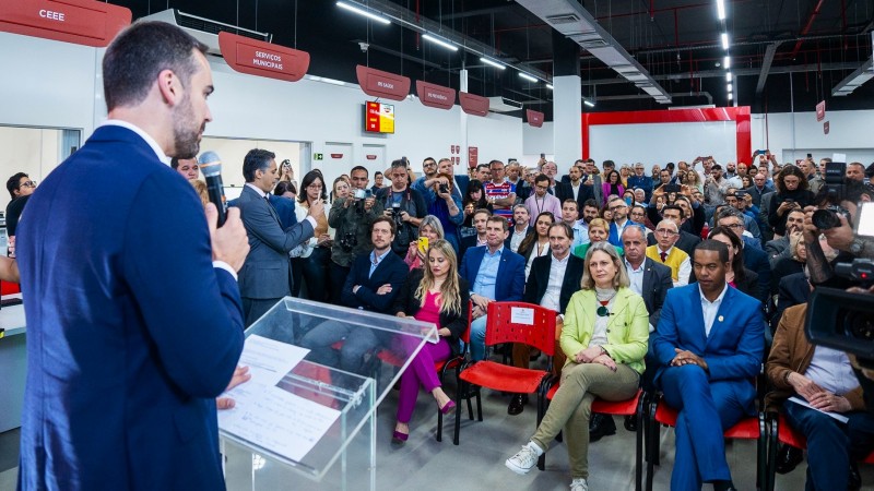 Leite de lado, ao microfone, do lado esquerdo da imagem. Ele está falando para uma plateia que pode ser vista à sua frentena inauguração do Tudo Fácil Pelotas.