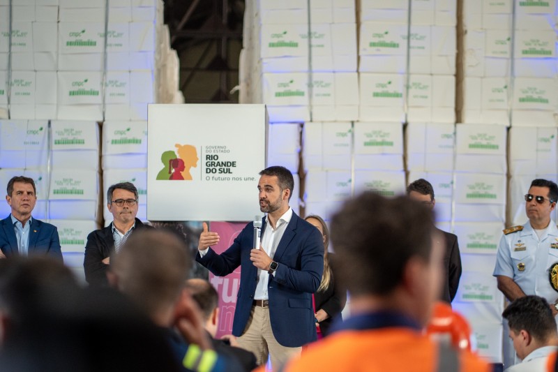 Contrato de dragagem do Porto do Rio Grande assinado nesta quinta (26) prevê investimento de R$ 94,5 milhões 2 Leite no centro da imagem. Ele está em pé, falando ao microfone dentro de um dos galpões do Porto do Rio Grande. Atrás dele são vistas quatro pessoas. Também podem ser vistas pessoas da plateia de costas assistindo ao governador. Contudo, essas pessoas estão desfocadas na imagem.