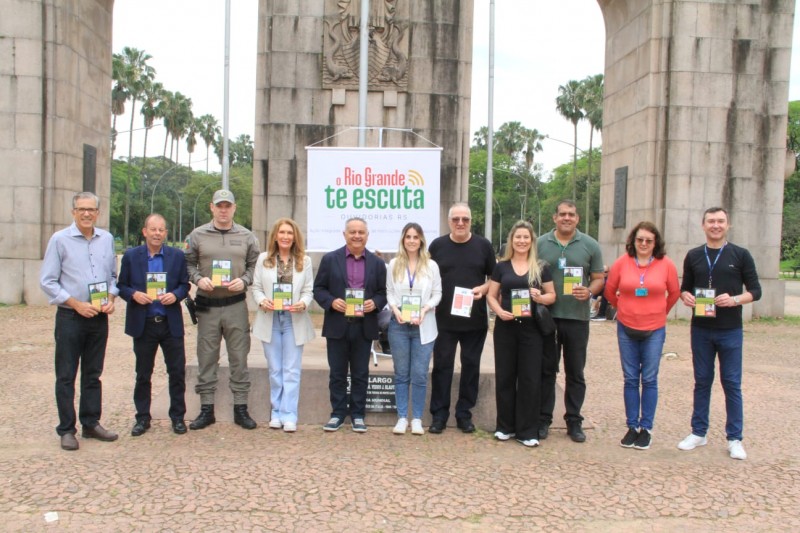 Ouvidores das instituições participantes do evento aparecem lado a lado em frente ao Monumento ao Expedicionário, no parque da Redenção, em Porto Alegre. Eles estão diante de um banner com a marca do projeto O Rio Grande te Escuta. Todos seguram nas mãos um exemplar do Guia das Ouvidorias.