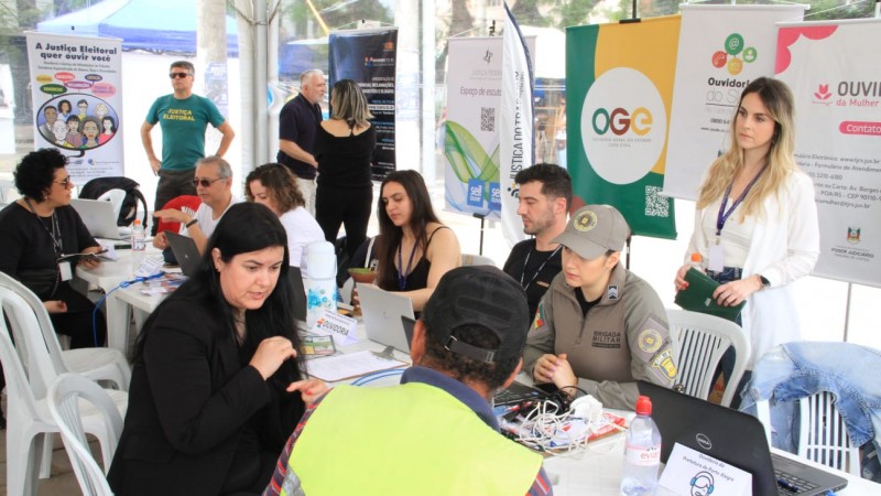 Servidores das ouvidorias participantes do evento aparecem sentados diante de mesas atendendo cidadãos que compareceram ao local. Ao fundo, estão banners com as logos das ouvidorias participantes.