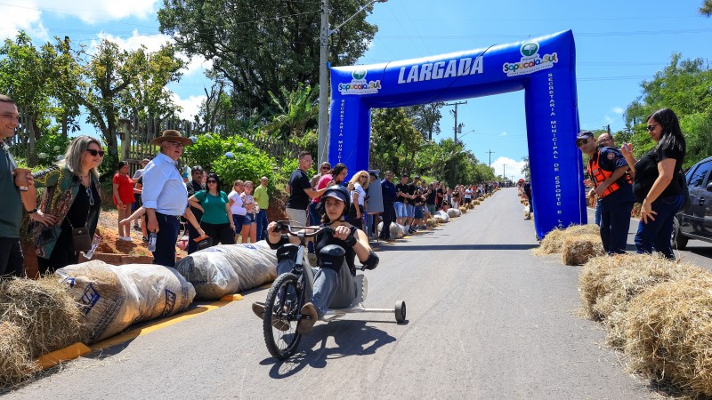 Uma mulher vestindo camiseta preta e calça jeans e tênis, e usando capacete e joelheiras, sentada em um carrinho de rolimã com guidon, desce uma lomba com a pista asfaltada. Ao fundo, um pórtico inflável azul sinalizada o ponto da largada. Ao lado da pista demarcada por sacos com feno, outras pessoas observam.