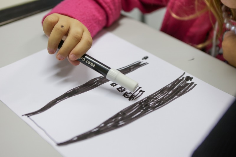Close em um desenho feito por uma criança e uma mão infantil que segura uma canetinha.