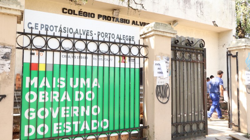 Imagem da fachada do Colégio Protásio Alves com destaque para uma placa com os dizeres Mais uma obra do Governo do Estado mais à esquerda da imagem, ao lado do portão de entrada.