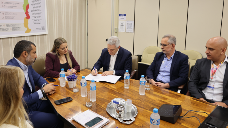 Záchia assinando o documento de posse na Procergs. junto com ele, à mesa de reunião, estão duas mulheres e um homem do lado esquerdo e dois homens do lado direito.