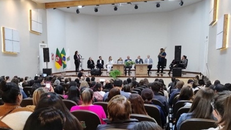 Imagem de um teatro. O público está de costas e em cima do palco há diversas pessoas sentadas e duas em pé durante algum momento da audiência realizada.