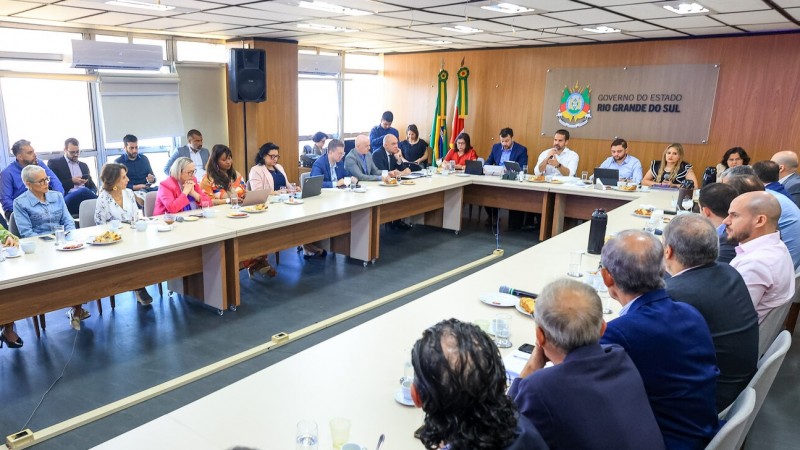Foto em ambiente fechado, em uma sala de reuniões. Ao centro, está o governador Eduardo Leite falando ao microfone, ao lado do vice Gabriel Souza. Os secretários estaduais estão sentados ao redor da mesa. 