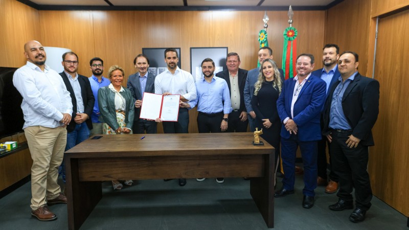 Foto em ambiente fechado. O governador Eduardo Leite está ao centro, em pé, segurando o documento da assinatura. Ao lado dele está o vice-governador Gabriel Souza. Ao redor deles, posando para a câmera, estão 11 autoridades.