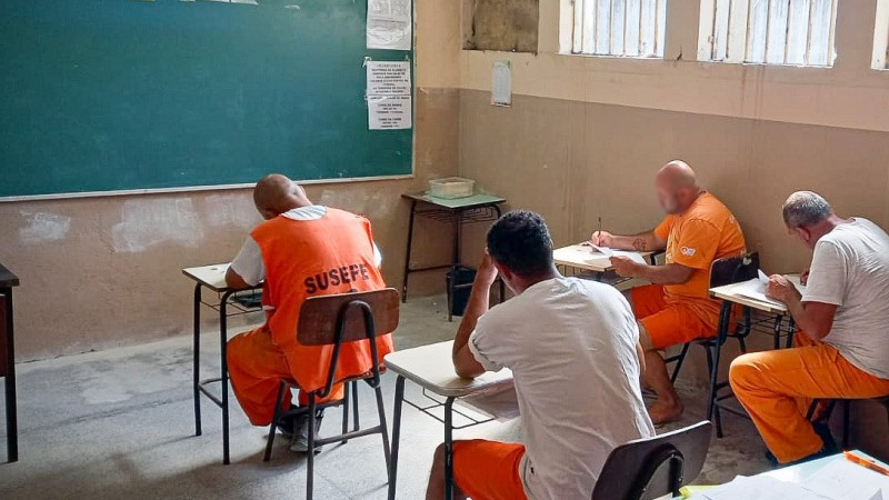 Foto em uma sala de aula. Há quatro apenados, vestidos com uniformes cor de laranja, sentados em mesas escolares e lendo materiais impressos. Na frente da sala, há um quadro verde.