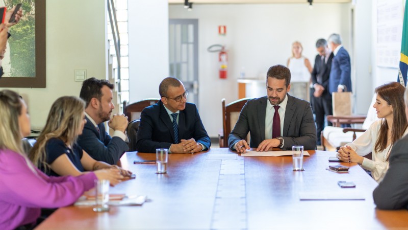 Em uma sala de reuniões, na ponta da mesa, estão o governador Eduardo Leite e o novo secretário de Habitação e Regularização Fundiária, que toma posse. Ao redor da mesa, acompanhando a cerimônia, estão outras autoridades. Todos estão vestidos em trajes formais. 