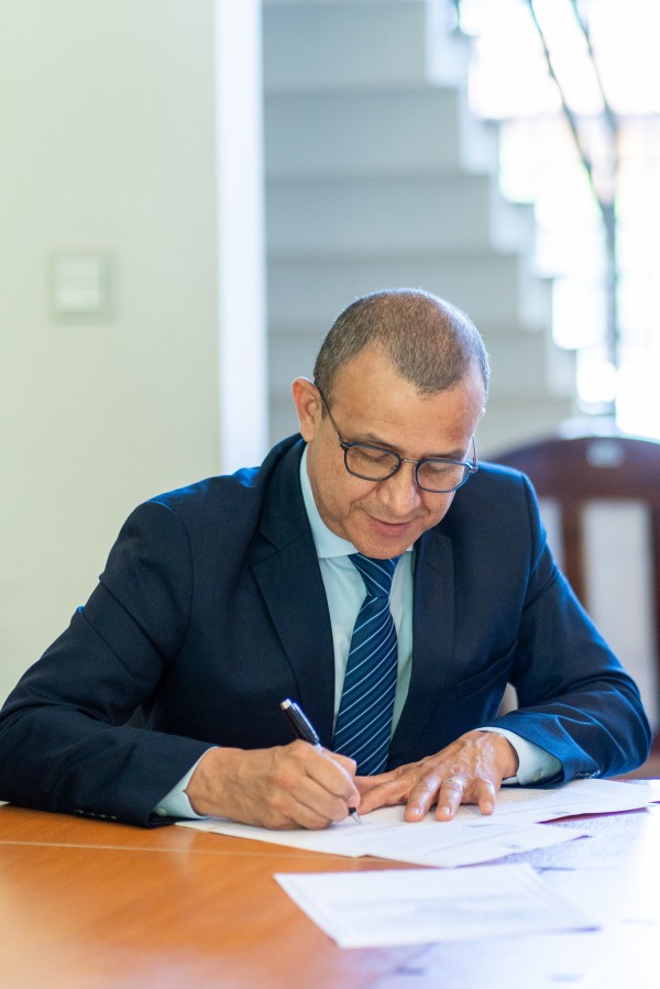Em uma sala de reuniões, um homem vestido com um terno azul marinho, cabelos escuros curtos e com óculos, assina o documento de posse. 