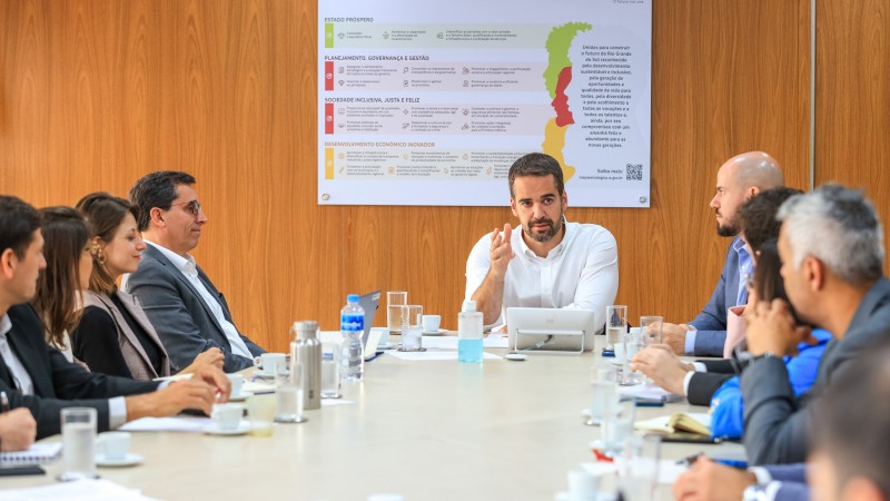 Em uma sala de reuniões, ao centro da imagem e na ponta da mesa está o governador Eduardo Leite, vestido com uma camisa branca, falando para as demais autoridades presentes na reunião, localizadas ao redor da mesa retangular. 