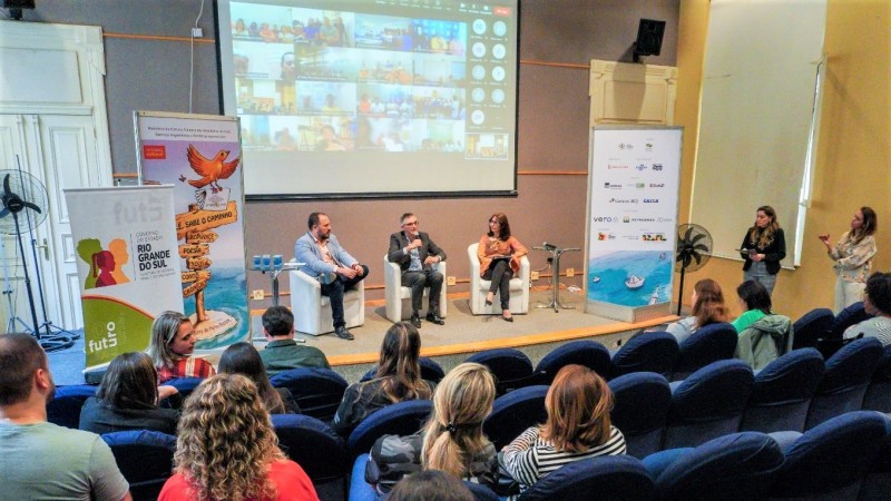 Imagem parcial de um auditório com três pessoas sentadas, em destaque, no palco, conduzindo o evento. A plateia é vista de costas. Ao fundo, há uma enorme tela que exibe várias janelas de uma ferramenta de teleconferência, nas quais são mostrados os participantes que estão em locais diferentes do local onde foi realizado ao evento.