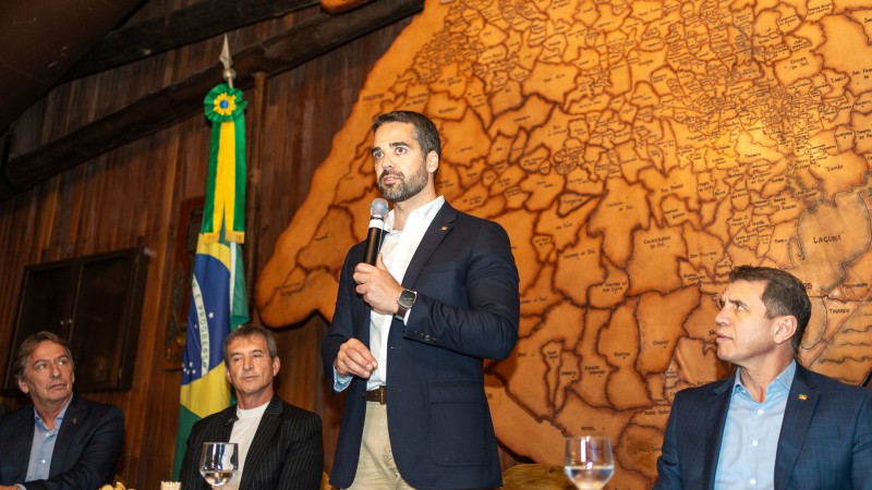Foto em ambiente interno. Leite está em pé, ao centro, falando ao microfone. Ao redor, sentados, estão três autoridades, olhando para o governador.