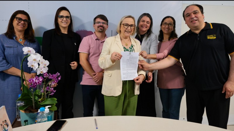 Foto em ambiente fechado. Cinco mulheres e dois homens posam para a foto, sorrindo. Todos est&atilde;o de p&eacute;. Ao centro, a secret&aacute;ria Arita segura o documento assinado. 