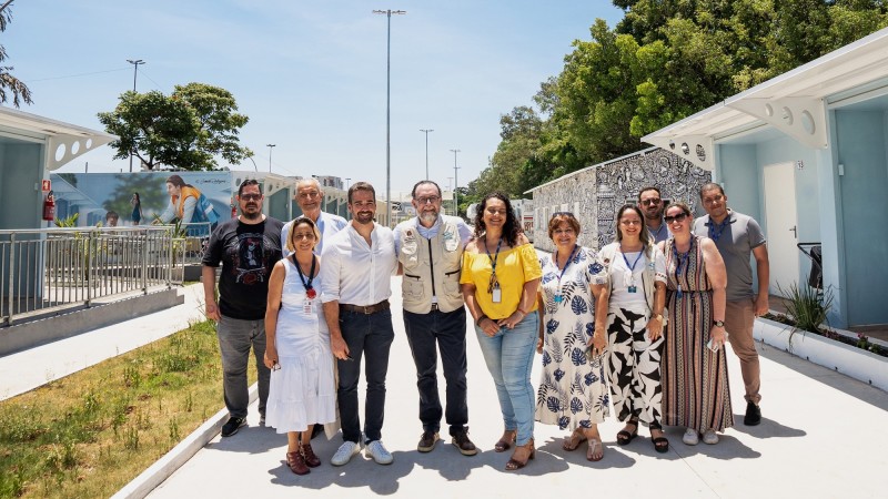 Foto posada. Todos em pé, Leite mais ao centro da imagem com uma mulher e dois homens à sua esquerda e quatro mulheres e dois homens à sua direita. Ambiente externo e aberto.