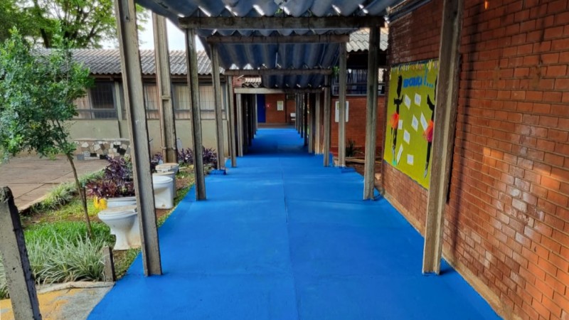 Foto do corredor de uma escola em obras. No lado direito da imagem, há uma parede de tijolos com um mural. No lado esquerdo, está o pátio da escola, com plantas e alguns vasos sanitários removidos.
