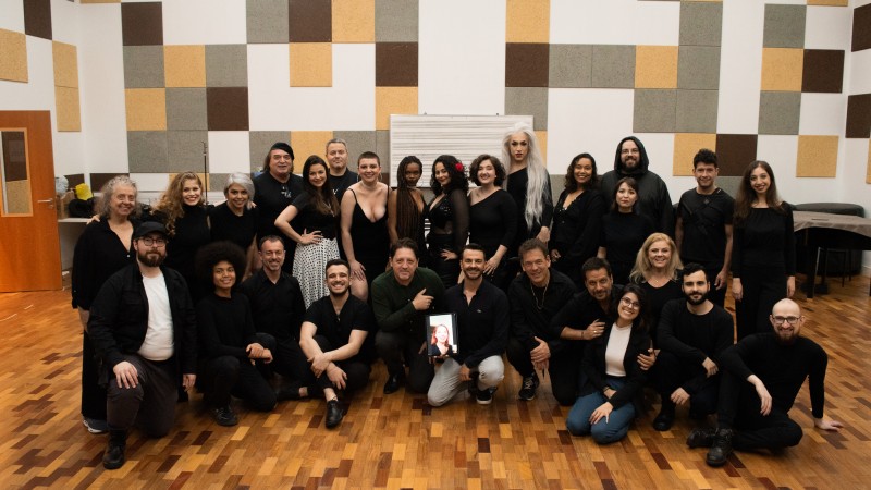 Foto posada em ambiente fechado. Em torno de 30 integrantes do espetáculo, a maioria em pé, e alguns agachados e sentados no chão, posa para a câmera trajando roupas escuras. A maioria sorri. O chão é um piso taco laminado.