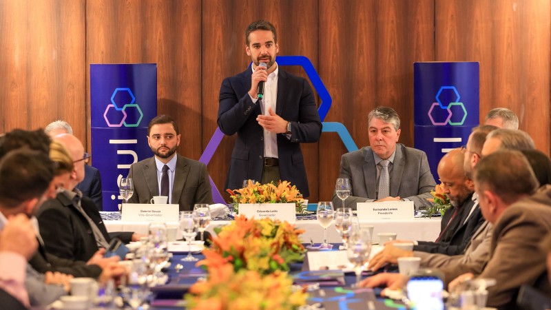 Leite em pé e falando ao microfone na cabeceira de uma grande mesa na qual são vistas pessoas do lado direito e do lado esquerdo e mais dois homens ao lado do governador. Atrás dele está o símbolo do Banrisul e dois totens nos quais está escrito Banrisul.