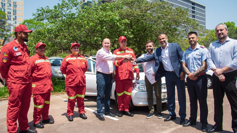 Foto posada em pé, com quatro bombeiros voluntários, sendo que um deles recebe simbolicamente as chaves de um automóvel doado das mãos do vice governador e de dois outros homens. Há mais dois homens à direita da foto participando da entrega.