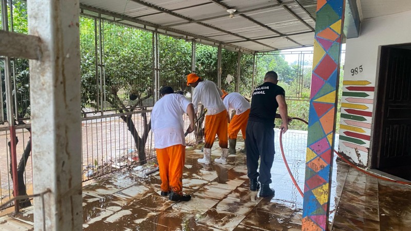 Apenados ajudando na limpeza de escolas em Arroio do Meio e Encantado após a enxurrada. Há um policial com a mangueira de água e três apenados limpando o chão com rodos e vassouras. Todos estão de costas na imagem.