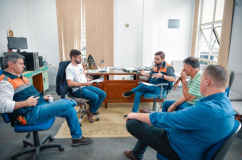 Em um sala, cinco homens estão sentados em cadeiras de escritórios numa roda. Há uma mesa ao fundo e outra à esquerda. No fundo, no centro, há uma janela coberta por persiana; há também uma janela aberta à direita. Na roda, Gabriel gesticula enquanto fala.