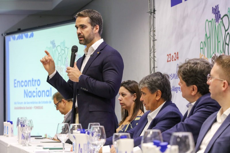 Leite ao microfone, em pé, gesticulando e falando para plateia. Três homens sentados estão à sua direita. Um homem e uma mulher podem ser vistos à sua esquerda.