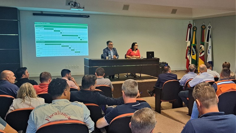 Um homem e uma mulher sentados atrás de uma mesa sobre um tablado falando para uma plateia. Atrás deles, mais à esquerda, há uma tela com uma tabela de cronograma sendo projetada.