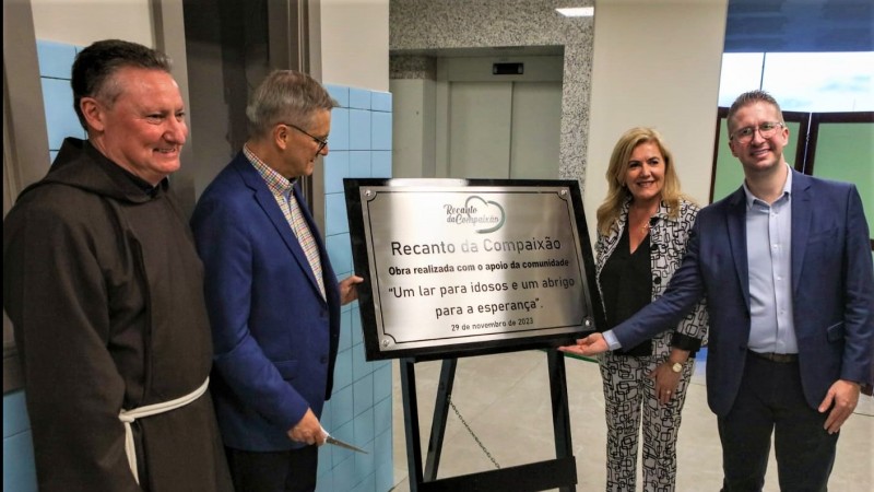 Dois homens apontam para uma placa que marca a inauguração do Recanto da Compaixão Frei Salvador em Caxias do Sul. A placa é relativamente grande e está colocada em um cavalete. Na imagem estão presentes o frei franciscano (vestido com túnica marrom) que irá cuidar do lar de idosos e uma mulher.