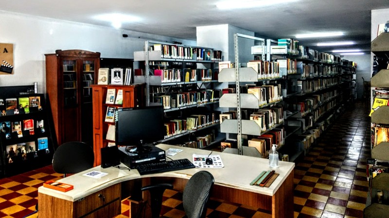 Foto interna mostra o Setor de Empréstimo da Biblioteca Pública Estadual, com diversas estantes cheias de livros, além de uma mesa com um computador em primeiro plano. 