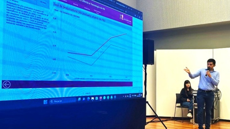 O diretor da Sict, em pé e ao microfone, mais à direita da imagem, apontando para uma tela à esquerda. Na tela, uma espécie de gráfico está sendo projetada.