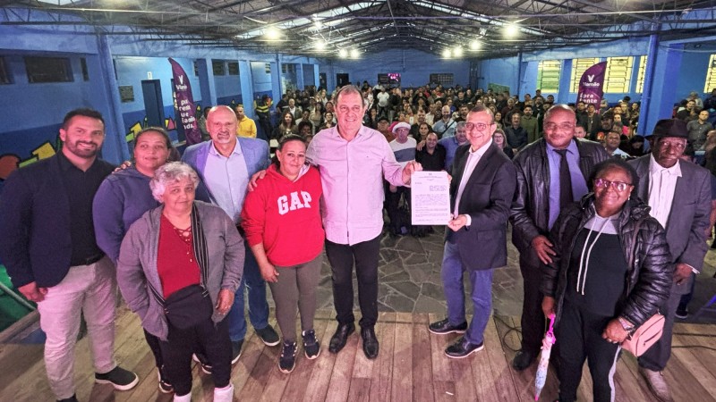 Foto posada. Dez pessoas em pé sobre um tablado e de costas para uma plateia grande. No centro o prefeito de Viamão e o secretário de Habitação e Regularização Fundiária seguram o documento de convênio que tinham acabado de assinar.
