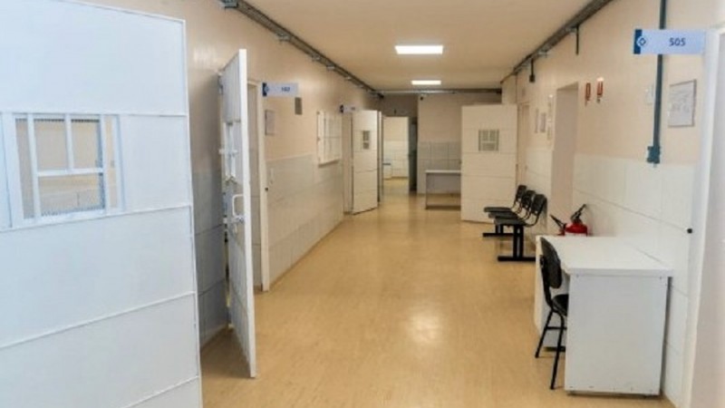 Imagem de uma ala hospitalar. Há um corredor com portas reforçadas abertas. Do lado direito podem ser vistas um escrivaninha com uma cadeira e mais ao fundo um conjunto de quatro cadeiras conectadas que parecem estra próxima de uma sala de atendimento.