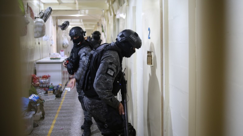 Foto tirada no interior de um presídio. A imagem enquadra um policial penal uniformizado com roupas escuras, com o rosto coberto, usando capacete e portando um rifle, olhando pelo vão da porta de uma cela, de cor creme. Ele está de lado para a câmera. Atrás dele, há outros policiais.